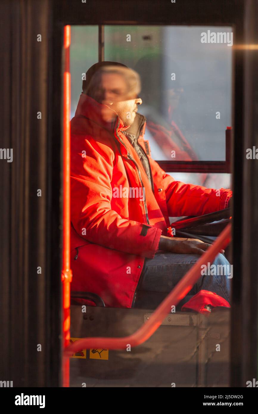 Bus driver dressed in a red coat. Red handrail. Face reflecting on driver's face. Brussels. Stock Photo