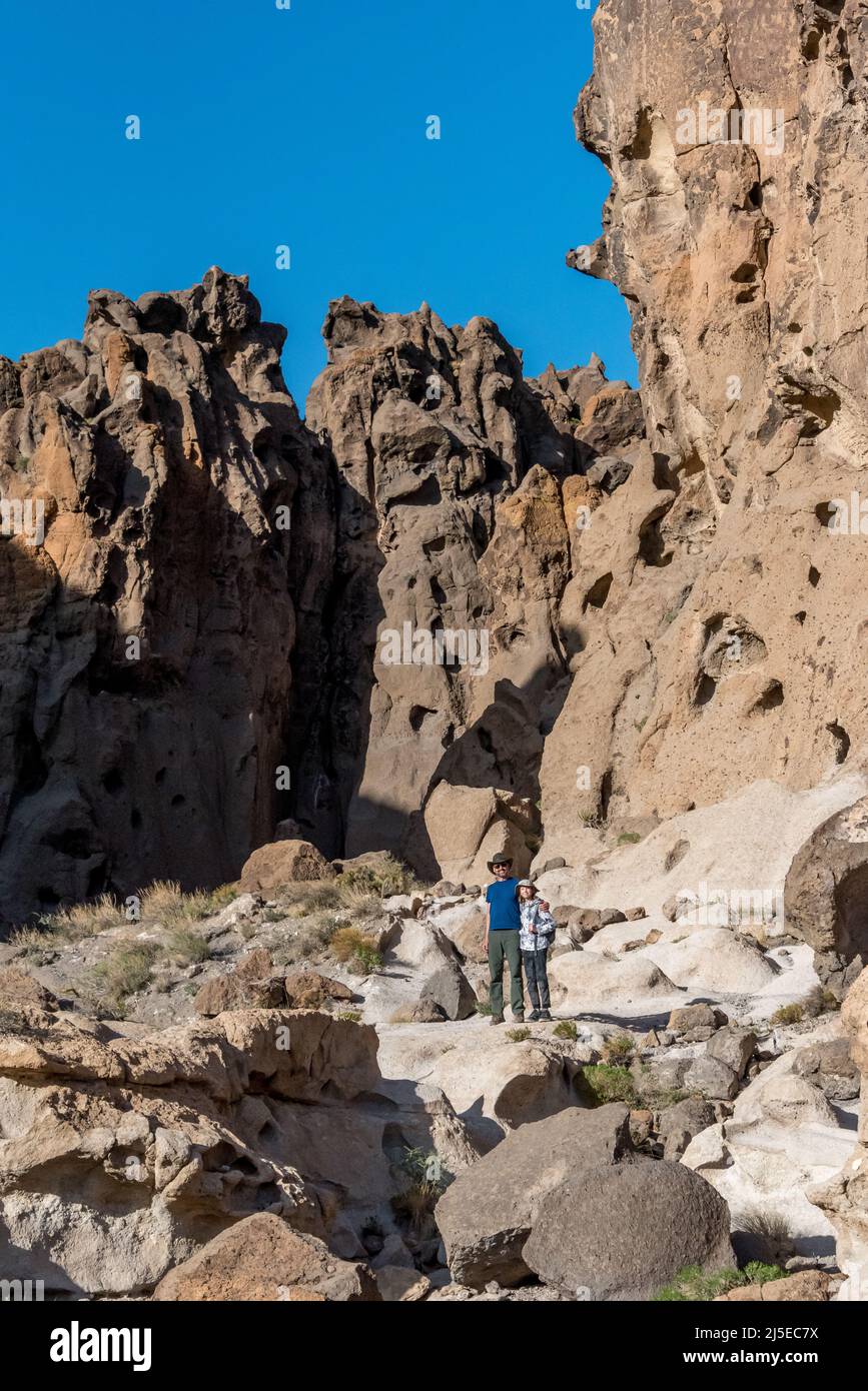 A father and son stand on boulders at Banshee Canyon with Hole in the Wall cliffs behind them, hiking Rings Loop trail in Mojave National Preserve. Stock Photo