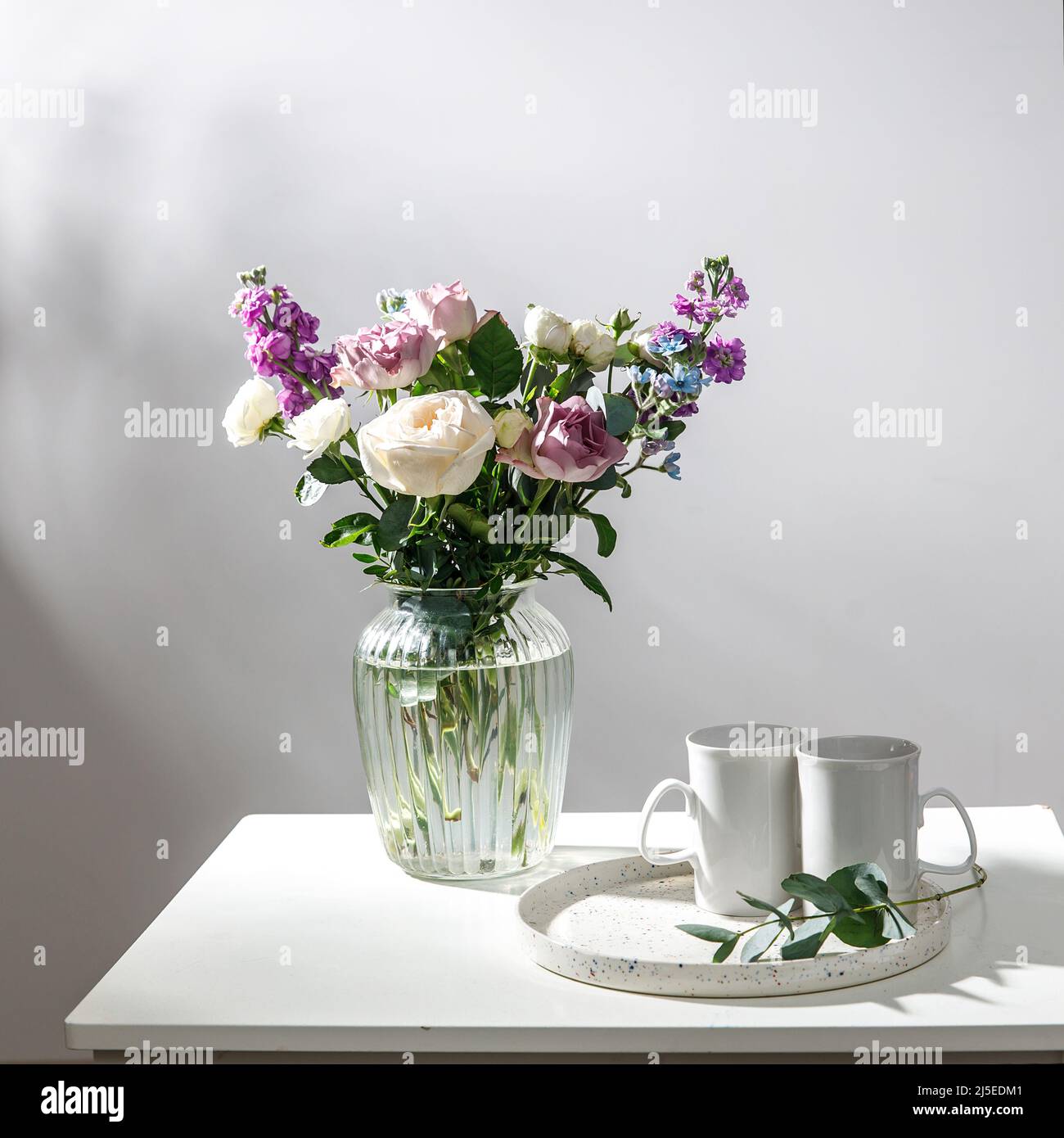 Bouquet of hackelia velutina, purple and white roses, small tea roses, matthiola incana and blue iris in glass vase is on the white coffee table with Stock Photo