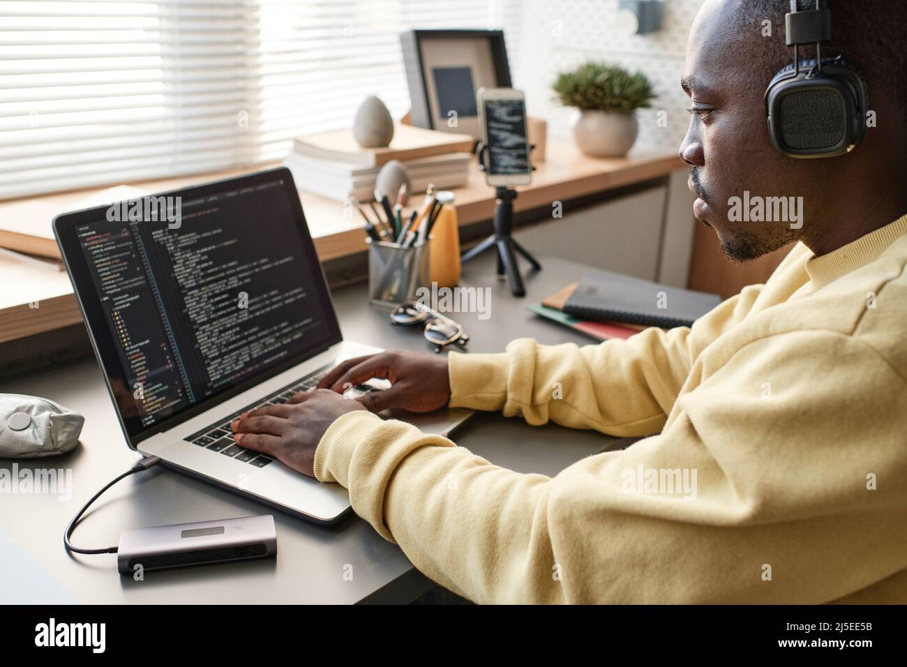Concentrated African American app developer in yellow sweater sitting at table and working on code using laptop Stock Photo