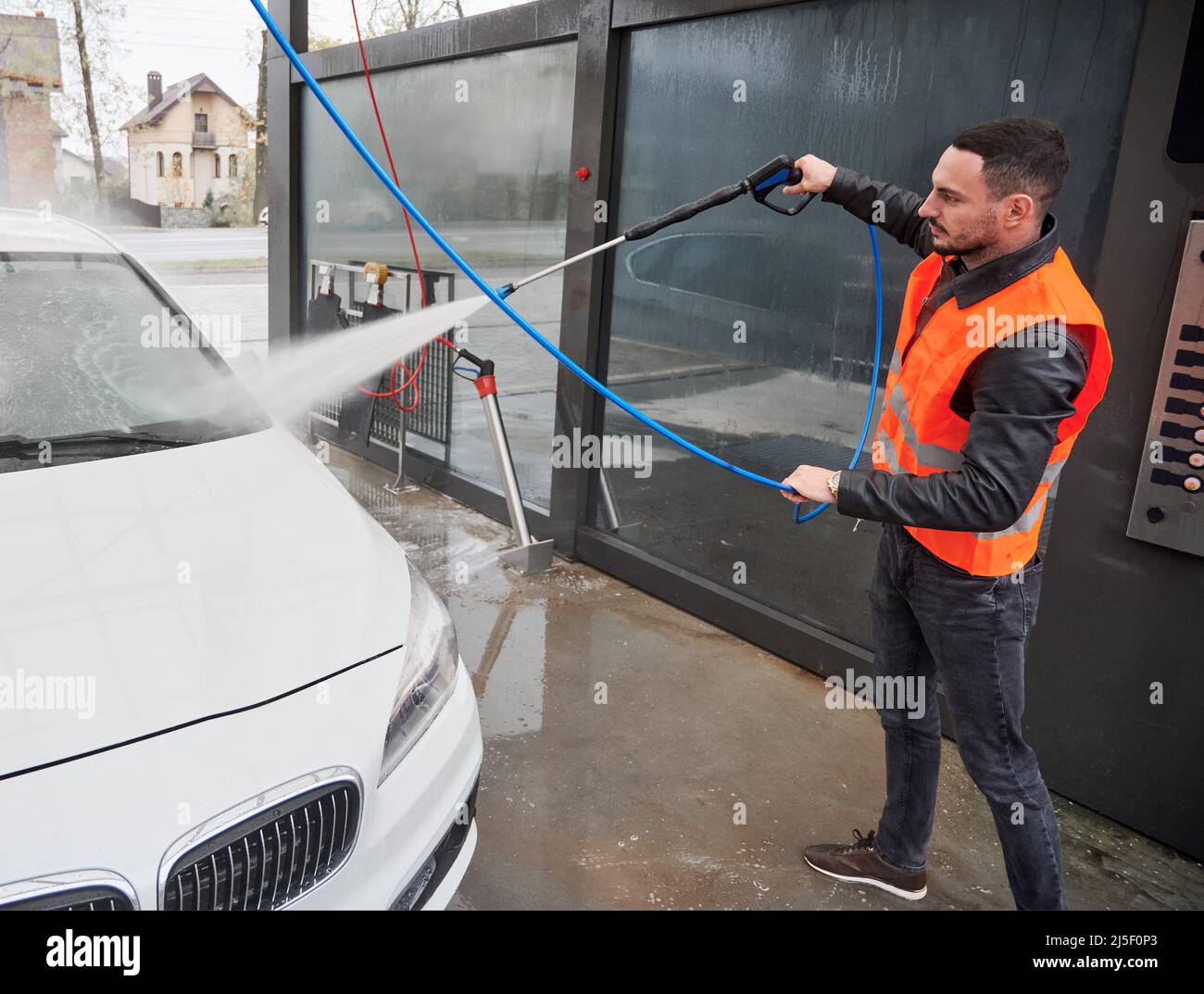 Young man washing white car on carwash station, wearing orange vest. Handsome worker cleaning automobile, using high pressure water. Stock Photo