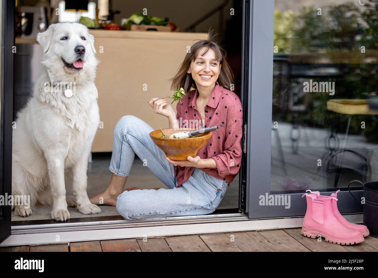 Happy woman eats salad while sitting with her adorable huge white dog at window Stock Photo