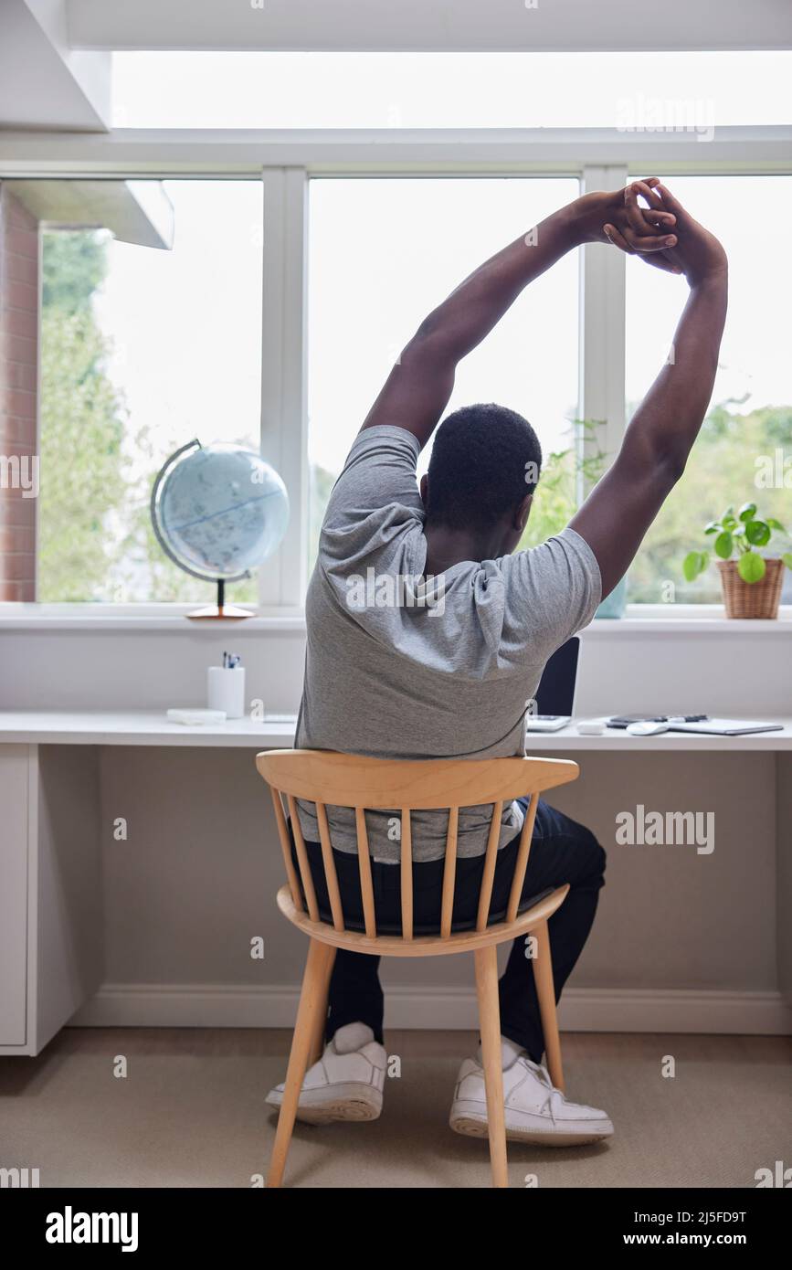 Rear View Of Man Working From Home On Computer  In Home Office Stretching At Desk Stock Photo