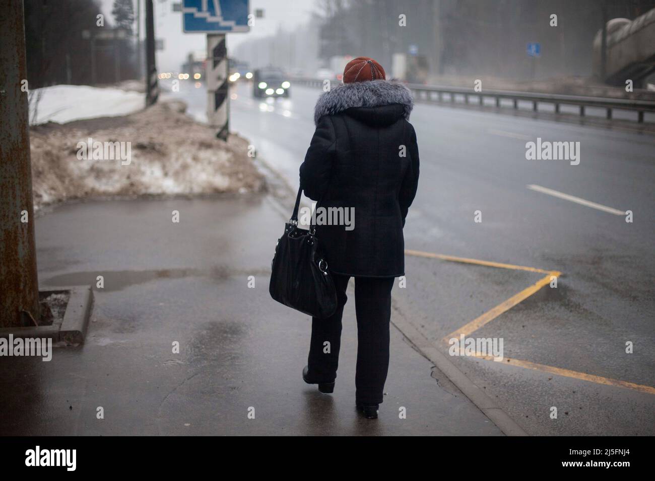 Woman is waiting for bus in Russia. Pensioner in bad weather stands by road. Girl in black clothes looks at highway. Stock Photo