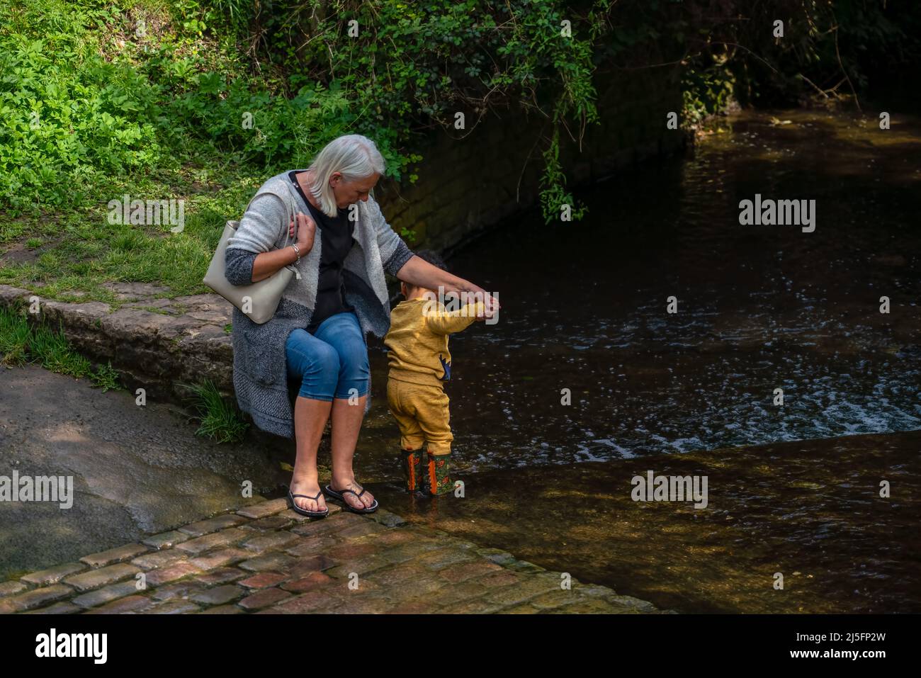 grandmother sits anmd holds the hand of her grandson as he paddles in a shallow brook, sunshine and dappled shade Stock Photo