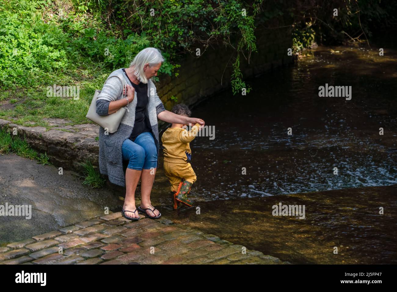 grandmother sits anmd holds the hand of her grandson as he paddles in a shallow brook, sunshine and dappled shade Stock Photo