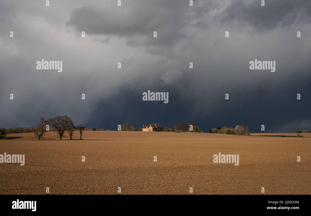 Dramatic stormy sky over a sunlit ploughed field with a farmouse in the distance. Stock Photo