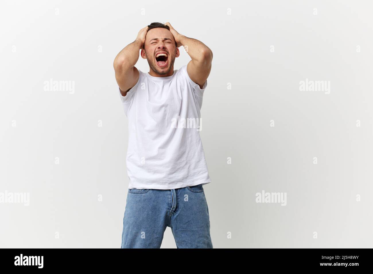 Overjoyed screaming angry tanned handsome man in basic t-shirt tearing his hair out posing isolated on over white studio background. Copy space Banner Stock Photo