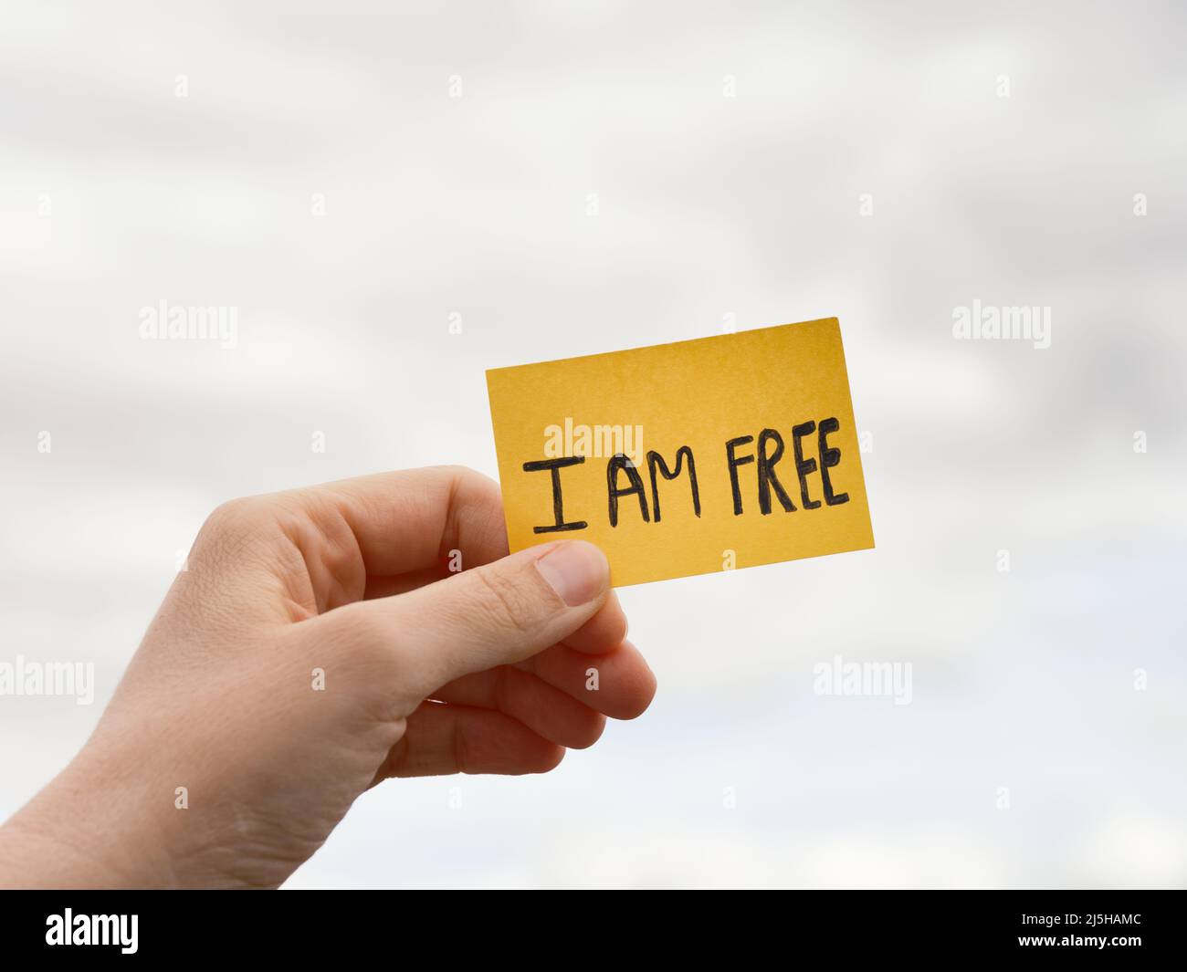 A woman holding a sticky note with the words I AM FREE over a overcast sky background. Close up. Stock Photo