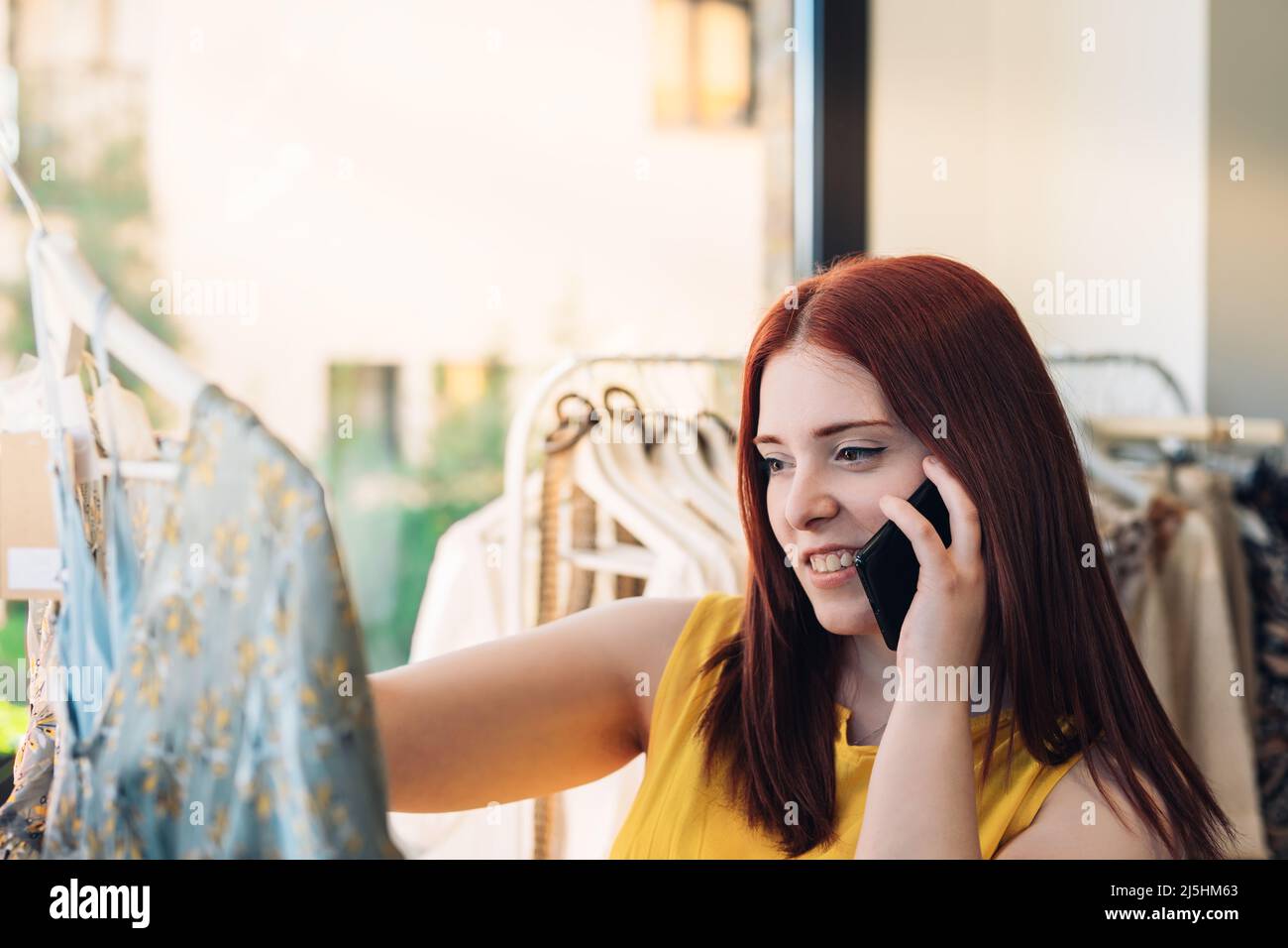 young red-haired woman shopping for clothes in a fashion shop. female talking on mobile phone. shopping concept. Stock Photo