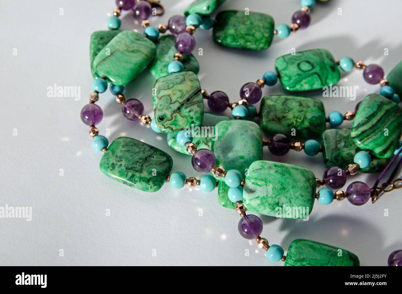 Coils of a handcrafted necklace made with beads of purple amethyst, blue turquoise and green crazy agate beads. Viewed on a white background with drop Stock Photo