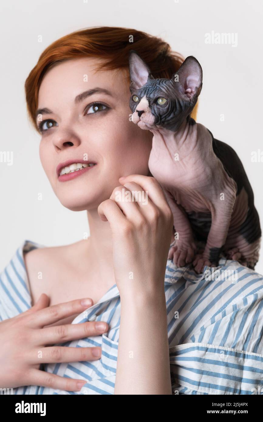 Sphynx kitten sitting on shoulder of happy redhead young woman. Selective focus on cat. Hipster female with short hair dressed in striped white-blue s Stock Photo