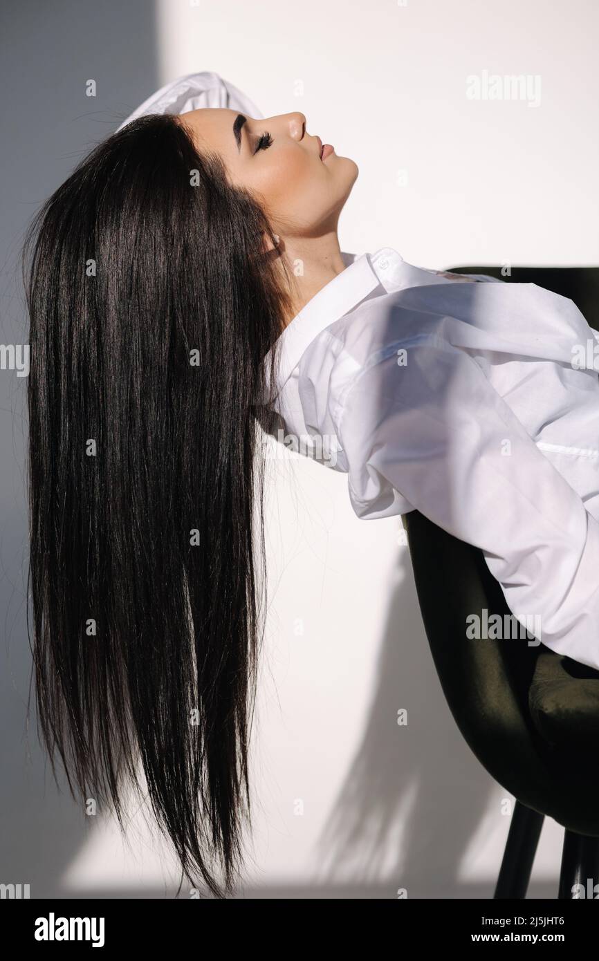 Female sitting on green chair. Her black hair is hanging down. Pretty woman in denim and white shirt Stock Photo