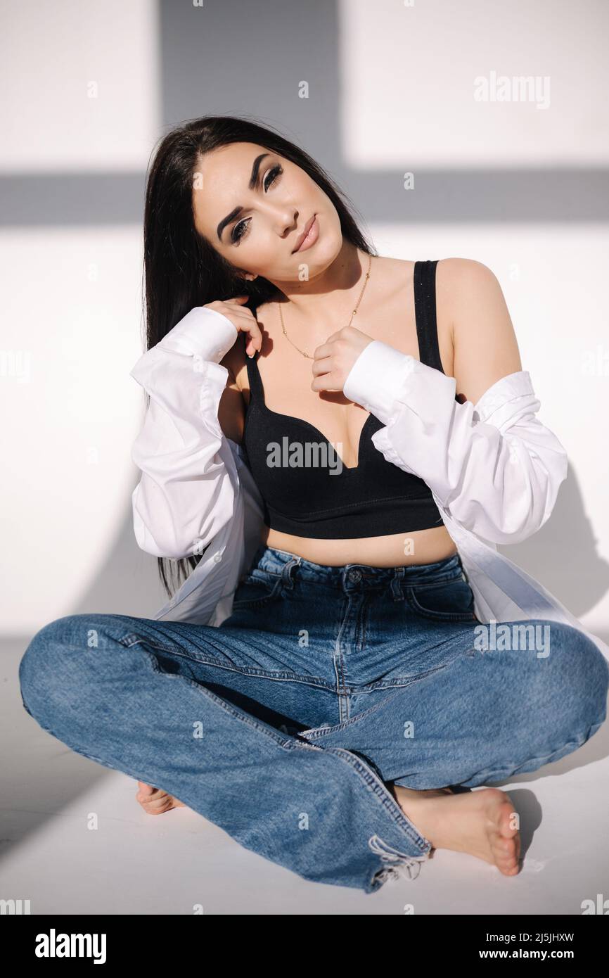 Close-up portrait of beautiful girl sitting on the floor in studio. Girl dressed in blue jeans and black topic and white shirt. Light from the sun Stock Photo