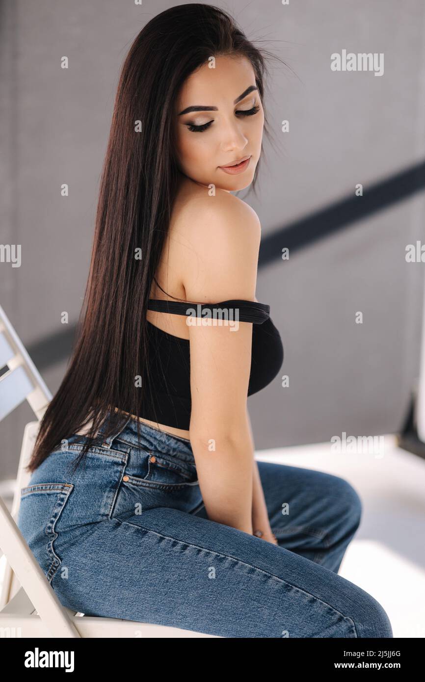 Sexy woman in black topic and blue jeans sits on chair during studio photoshoots. Brunette girl posing to photographer. Shadow on white background Stock Photo