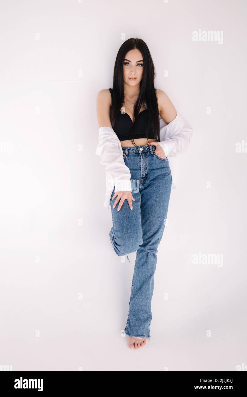Pretty woman barefoot in studio. Beautiful brunette girl in black topic with white shirt and blue denim jeans. White background of cyclorama Stock Photo