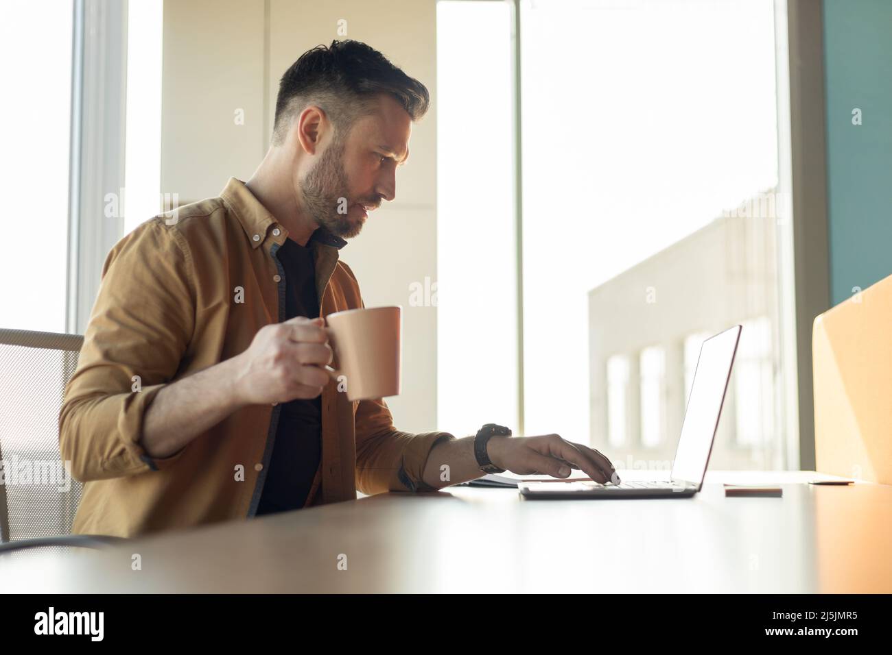 Man Working On Laptop Drinking Coffee In Office, Side View Stock Photo