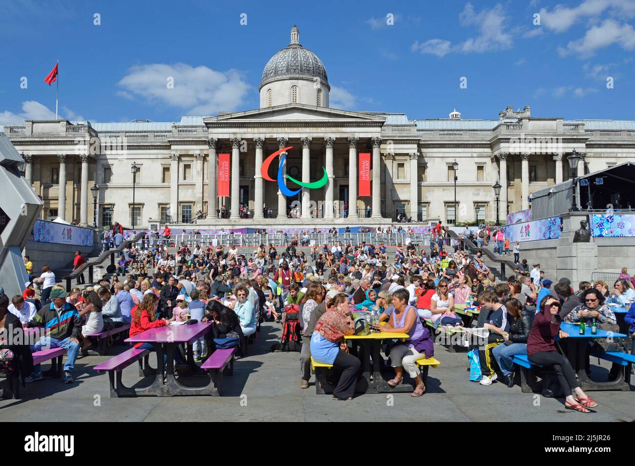 Tourists & visitors sit at coloured picnic tables in Trafalgar Square 2012 London Paralympic Games logo on colonnade of National Gallery England UK Stock Photo