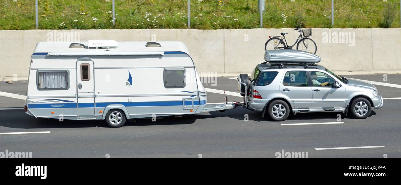 Man driving silver grey car fitted with roof rack top box supporting ladies bike with shopping basket towing white caravan driving on UK motorway road Stock Photo