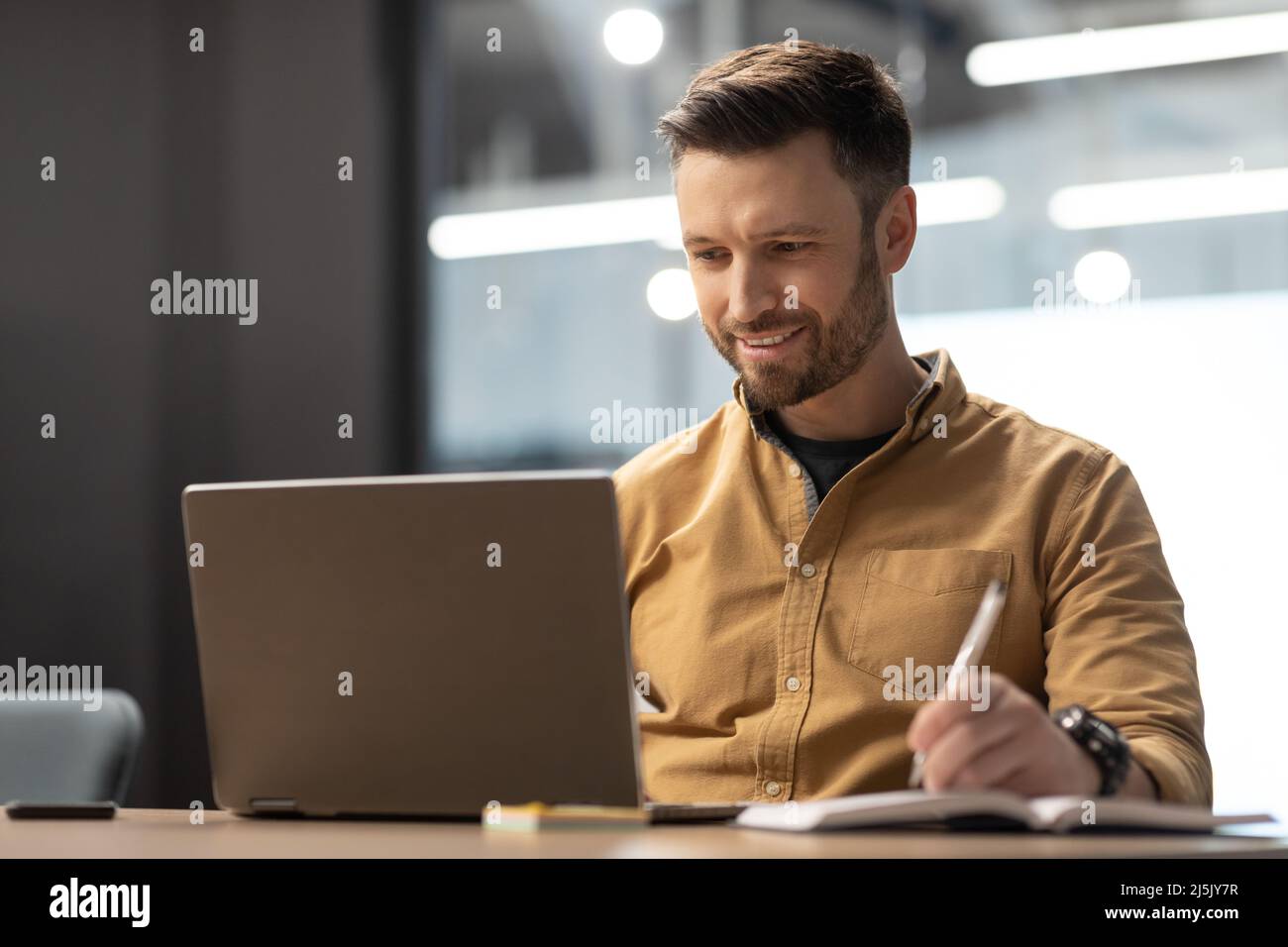 Cheerful Entrepreneur Using Laptop And Taking Notes Working At Workplace Stock Photo