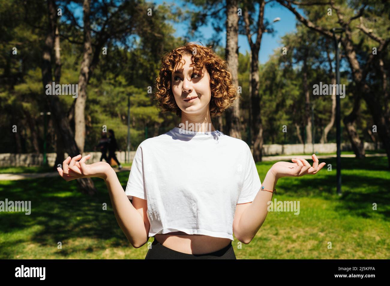 Unsure woman wearing white tees and black yoga pants shrugging shoulders over at outdoor. Young redhead woman looking away. Hesitating woman. Helpless Stock Photo