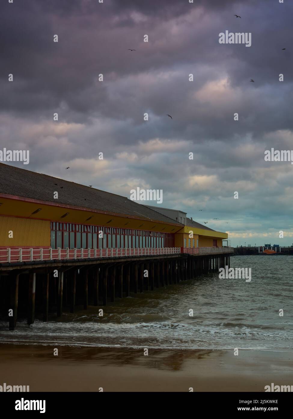 A fun fair complex on a pier, extending out into the sea at Frinton. Seagulls circle overhead in a storm-clouded sky as waves crash around the columns Stock Photo