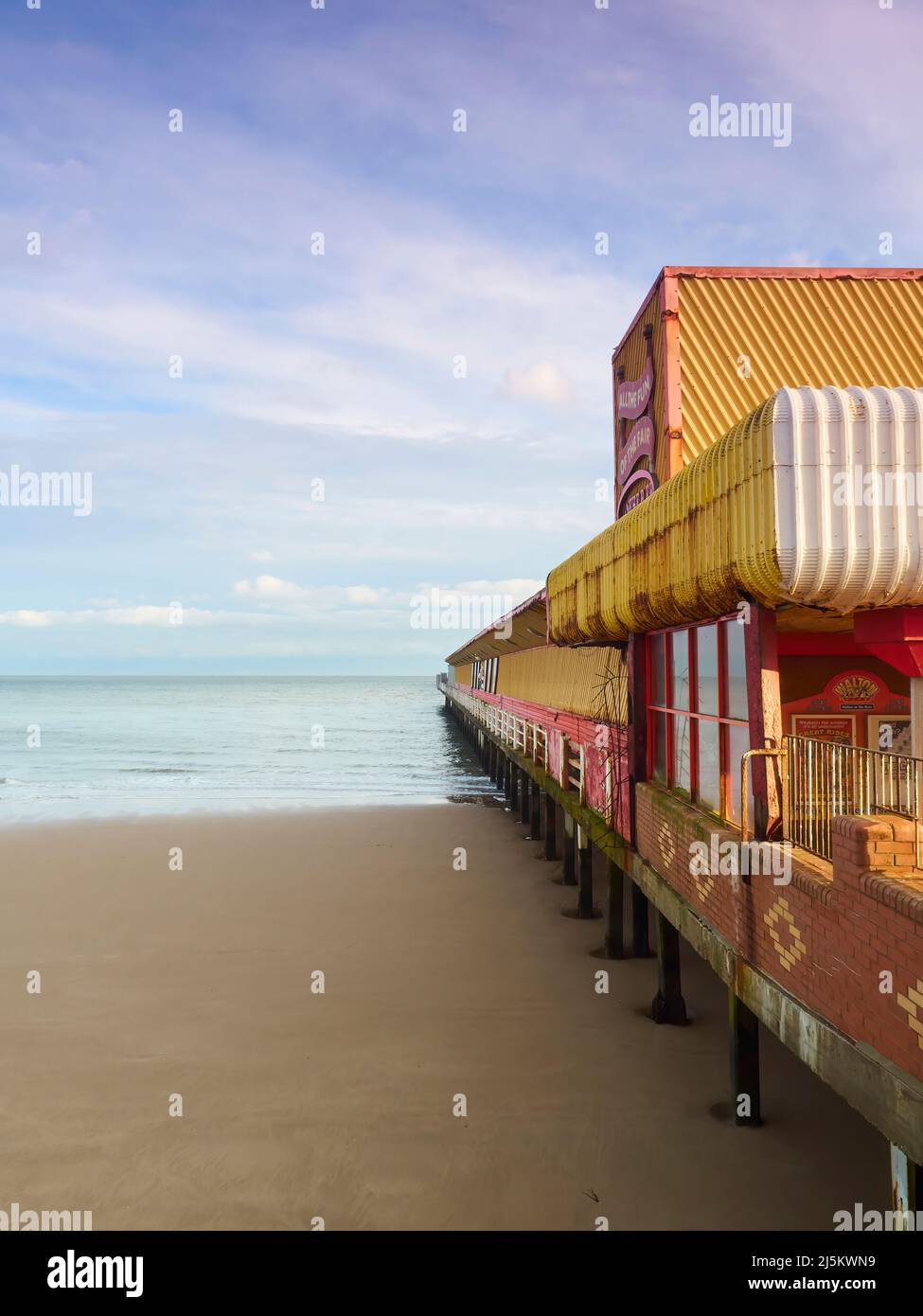 A fun fair complex on a pier, extending out into the sea at Frinton. Soft sunlight and wispy clouds give a wistful, nostalgic feel. Stock Photo