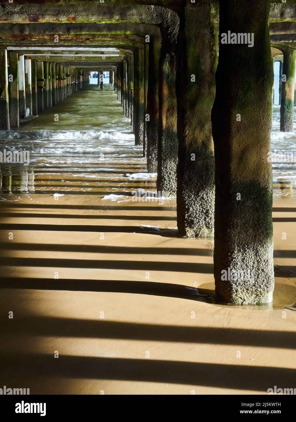 Underneath the fun-fair at Frinton at low tide, slanting sunlight casting sharp stripes of shadows on the wet sand as waves rollback in. Stock Photo