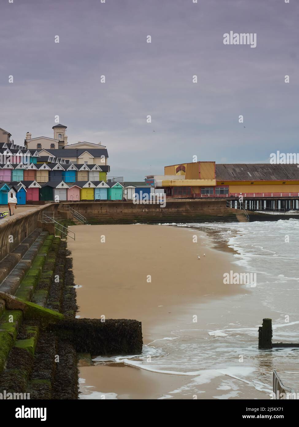 Colourful beach huts on a hill curving to a derelict looking fun fair, wrapping around a bay of breaking waves and sea defences. Stock Photo