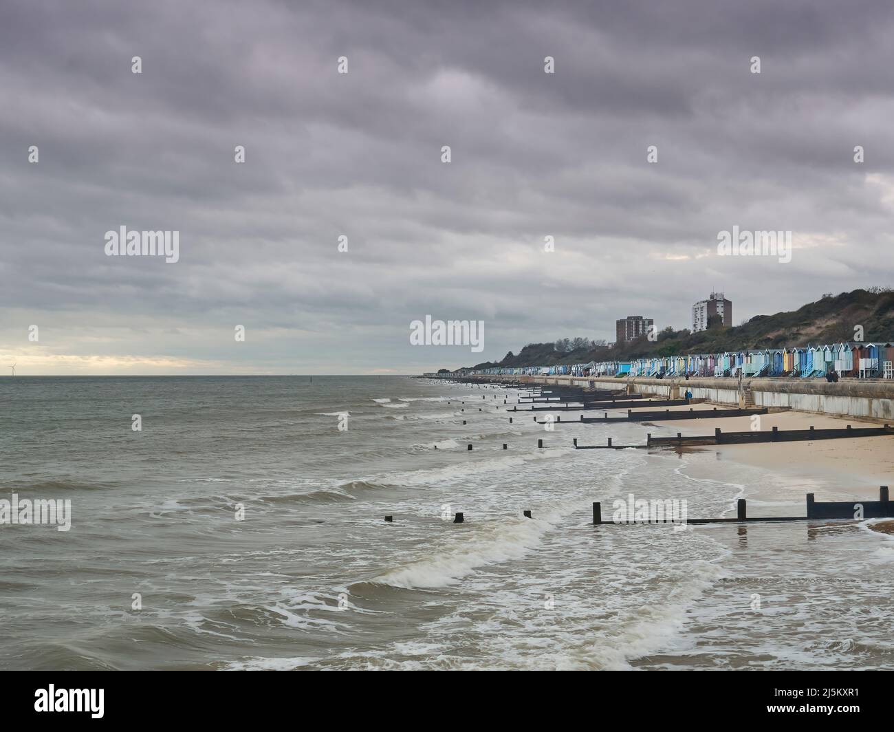 A sweep of beach, sea defences, beach huts and hills at Frinton, standing against breaking waves under a stormy sky with a tear admitting sunlight. Stock Photo
