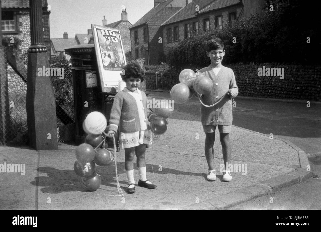 circa 1938, historical, two young children standing on the corner of a village square selling balloons to raise funds for the Shipwrecked Mariners Society, England, UK. Founded as long ago as 1839, and still operating today, the charity provides support to the retired merchant seafarers and fisherman and of course those affected by shipwrecks. Beside the letterbox, a poster for the  'flag & baloon' day for 'The Shipwrecked Mariners' Society', note the different spelling of balloon. Stock Photo