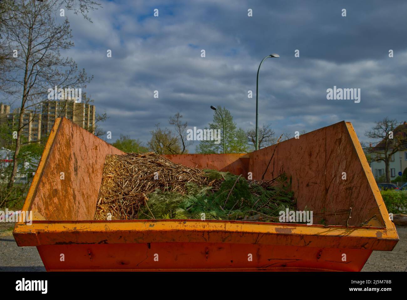 big orange-colored steel waste container with green waste in contrast to a dark clouded sky in grey and blue Stock Photo