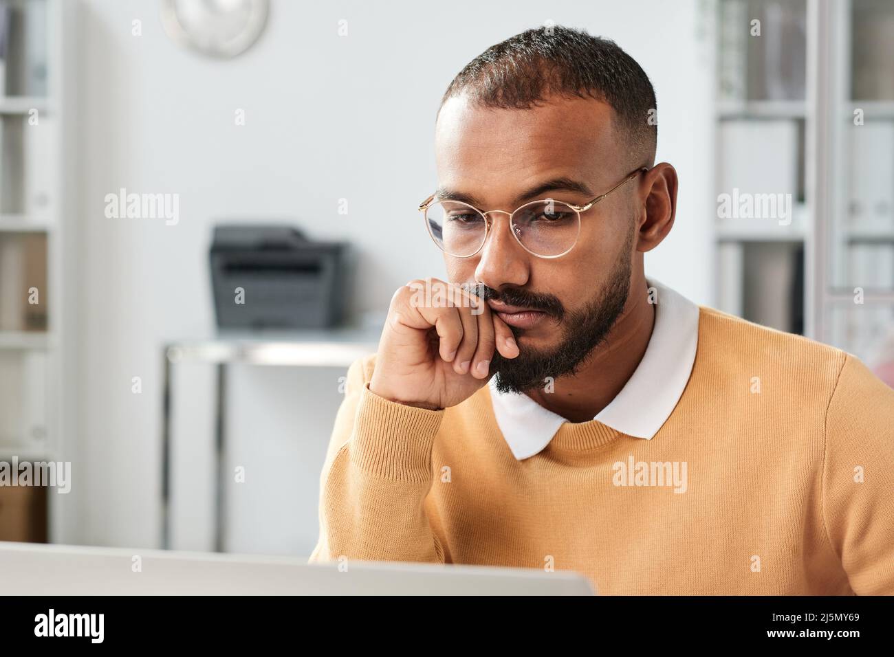 Pensive young middle eastern man in eyeglasses leaning head on hand while working with analytics in office Stock Photo