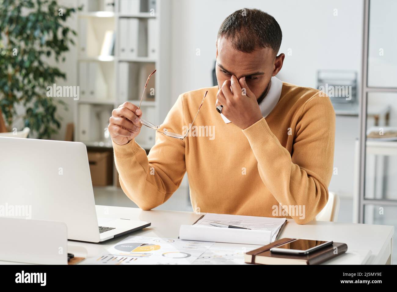 Tired young middle eastern businessman holding eyeglasses and rubbing bridge of nose while overworking in office Stock Photo