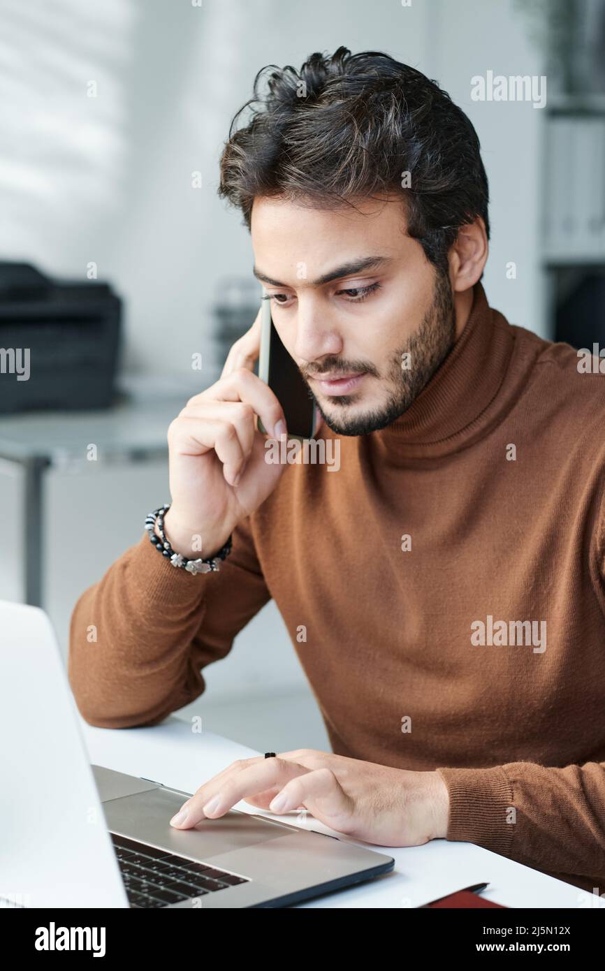 Serious modern businessman in brown turtleneck sitting at table and using laptop while having phone conversation Stock Photo