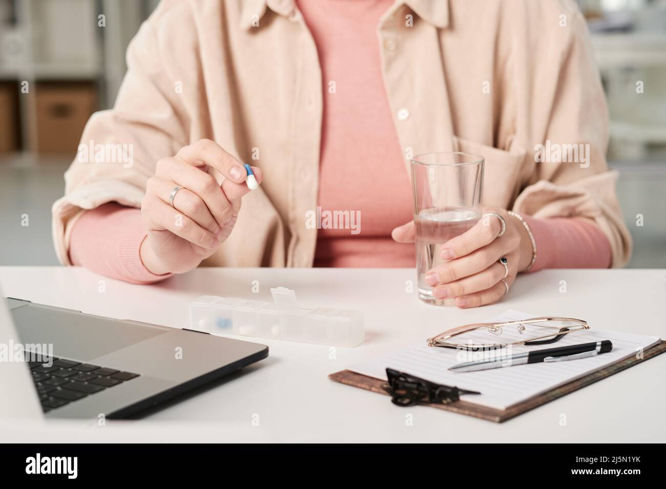 Close-up of unrecognizable woman sitting at office table with pill organizer and glass and taking biohacking supplement Stock Photo