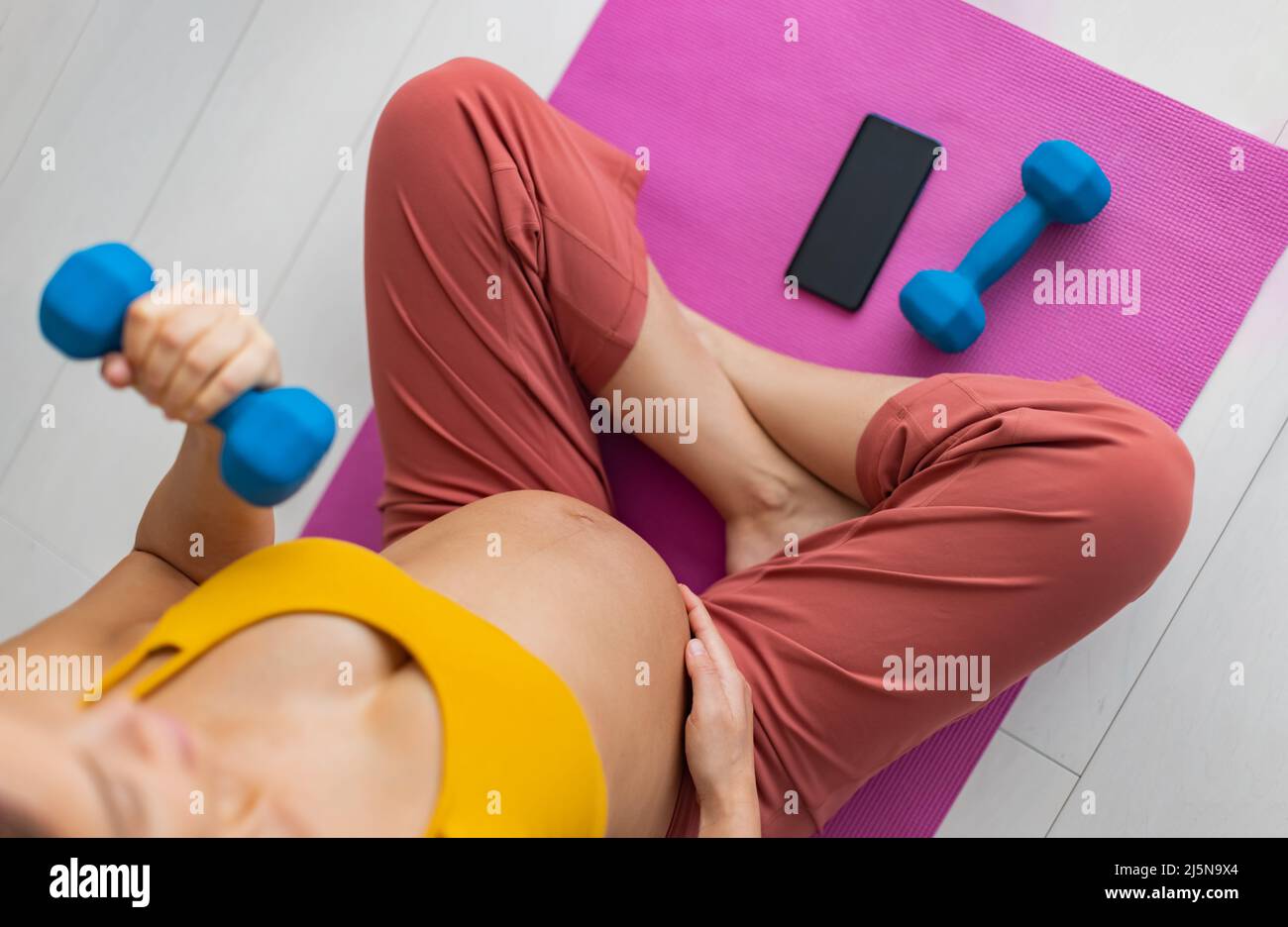 Pregnant woman working out at home training with online fitness phone app lifting free weight for arm workout Stock Photo
