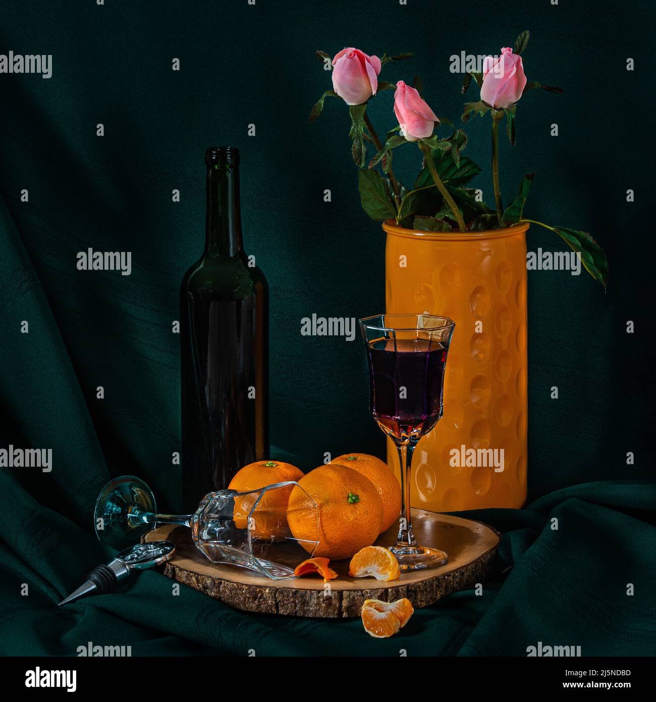 A dark and atmospheric photo of a still life, a bottle of wine, glasses, oranges and a bottle with roses. Stock Photo