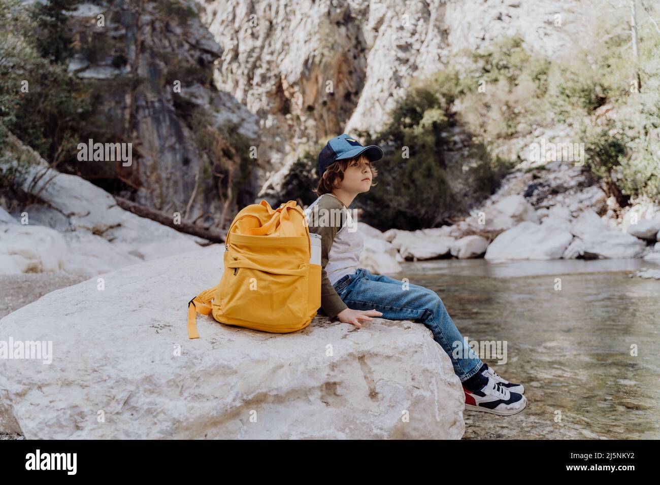 School boy with yellow backpack sits on a riverside rock in the canyon with mountain cliffs in the background. Kid child taking a rest on a boulder Stock Photo