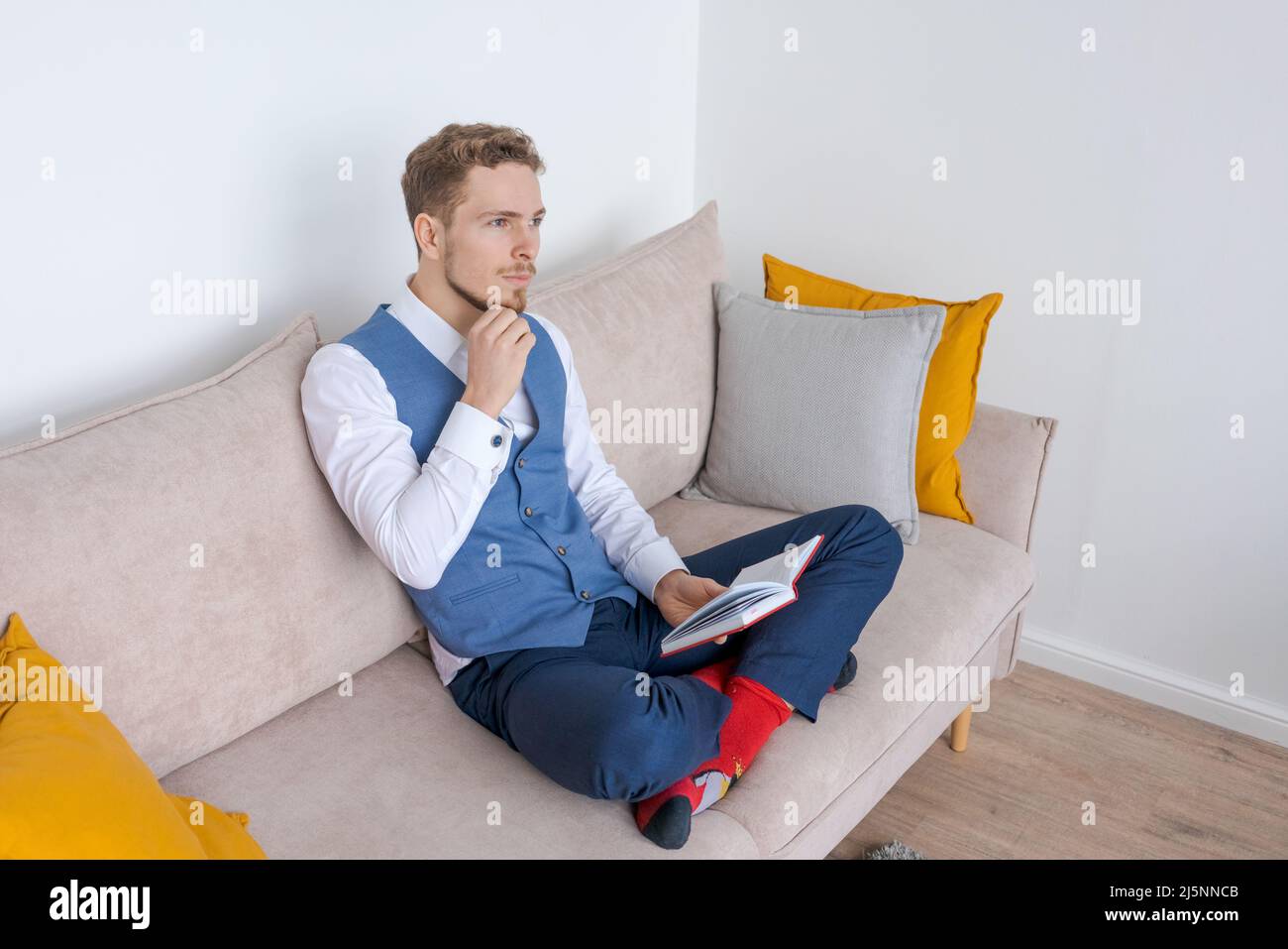 Pensive businessman sits on sofa with diary in his hand, thinks about plans in a blue bearded guy suit. Plans his daily routine and business meetings. Record keeping and planning concept Stock Photo