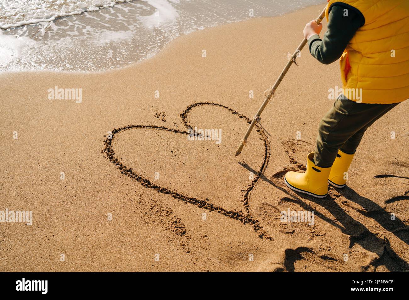 Boy in yellow rubber boots drawing heart shape on sand at the beach. School kid touching water at autumn winter sea. Child having fun with waves at Stock Photo