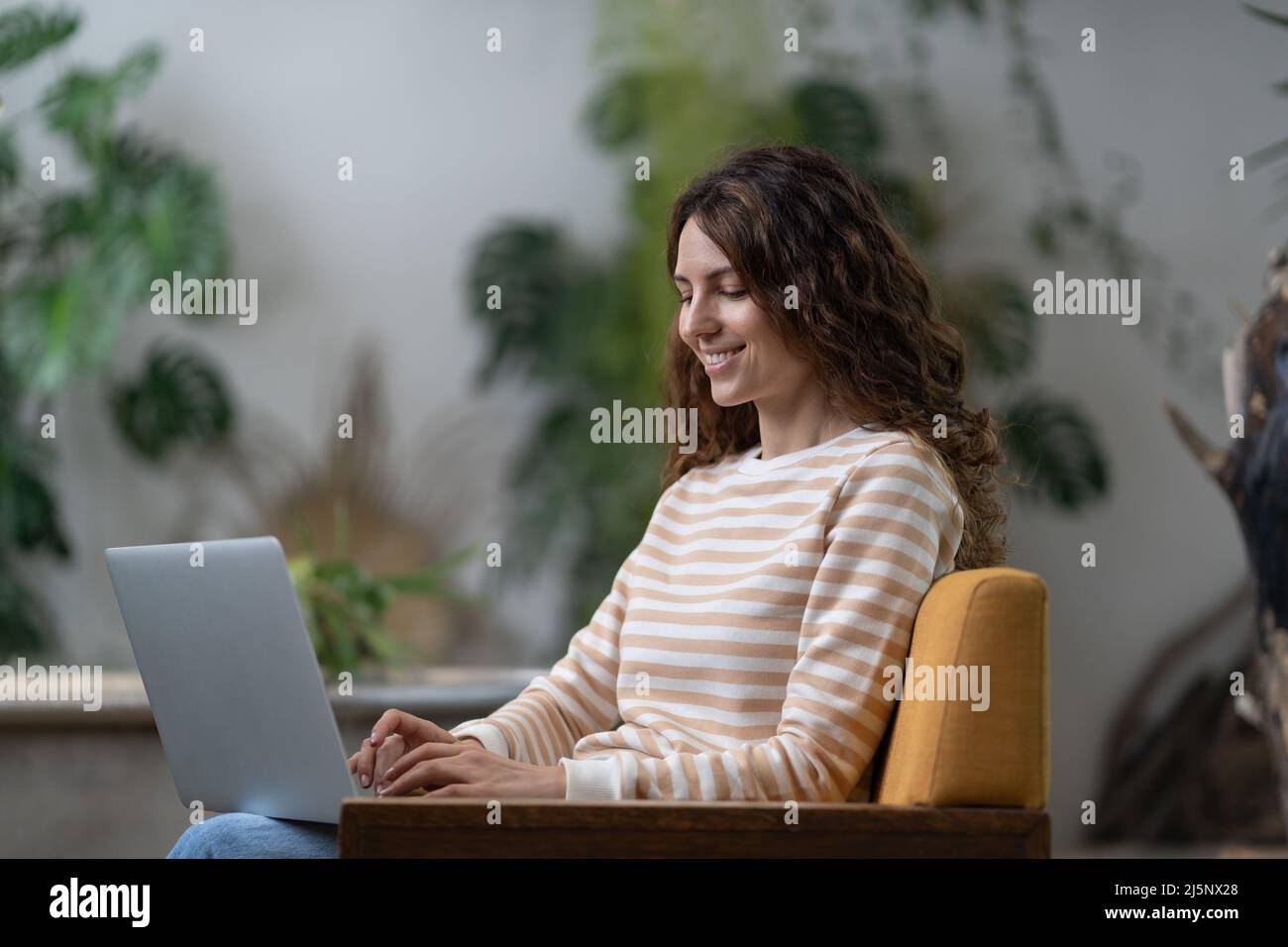 Freelancer young woman sitting in armchair in greenhouse, working on laptop surrounded by houseplant Stock Photo