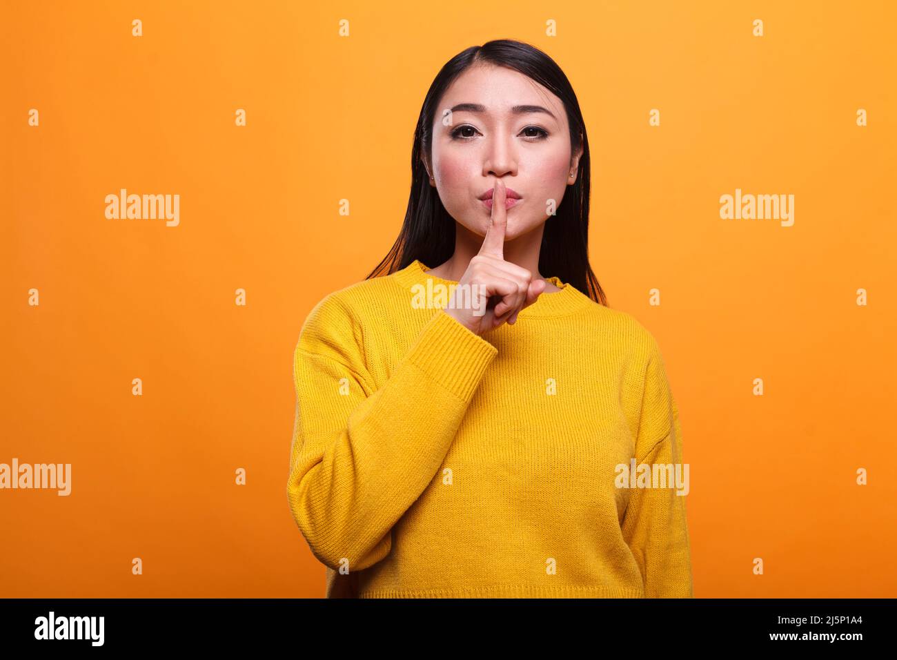 Beautiful mysterious woman wearing vibrant yellow sweater making silence gesture on orange background. Secretive person touching lips with forefinger indicating silence and secrecy. Stock Photo