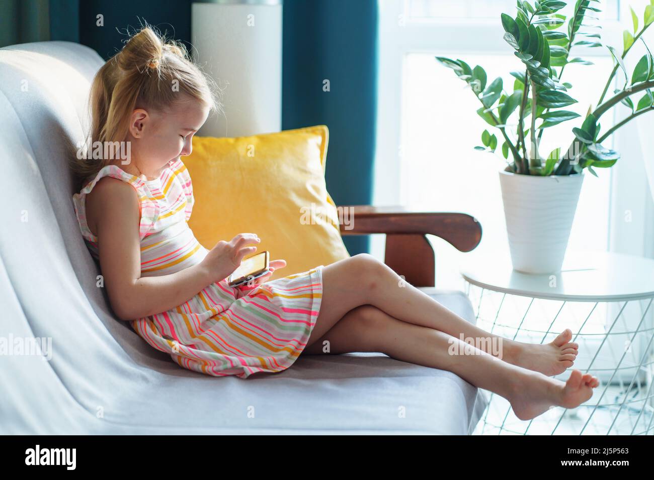 Schoolgirl in casual dress using smartphone on sofa in living room side view. Stock Photo