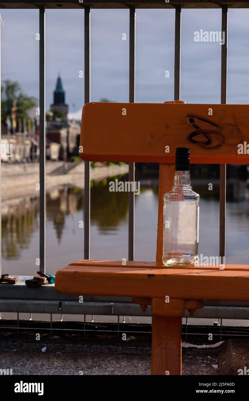 Empty alcohol bottle on a bench alongside the river with blurred city and church tower in the background Stock Photo