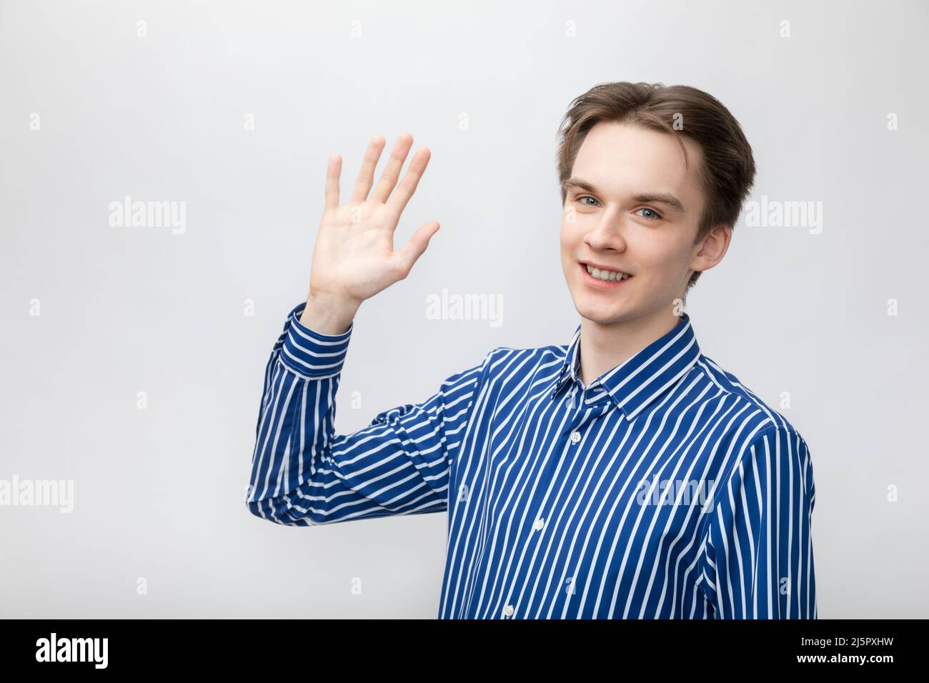 Portrait of happy cheerful young man wearing blue-white striped button shirt waving with hand saying hi looking at camera smiling. Studio shot on gray Stock Photo