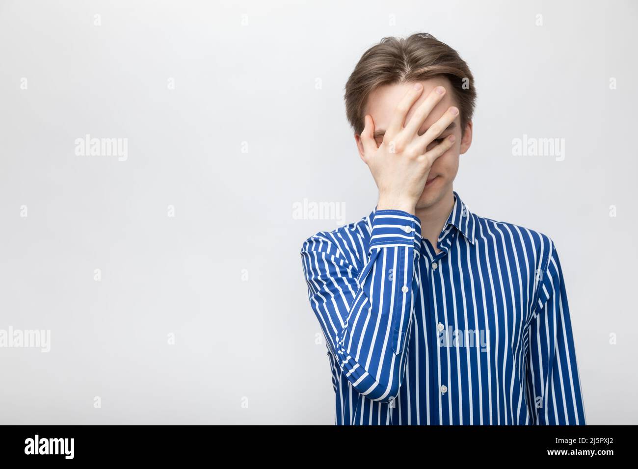Portrait of young man wearing blue-white striped button shirt covering his eyes and face with palm. Studio shot on gray background Stock Photo