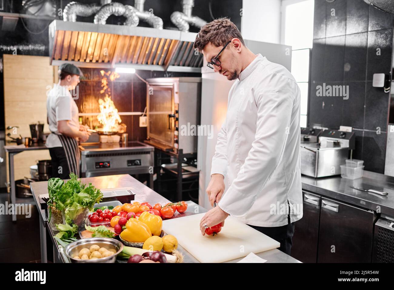 Young cook in uniform cutting vegetable on cutting board at table while his colleague using open fire to fry them Stock Photo