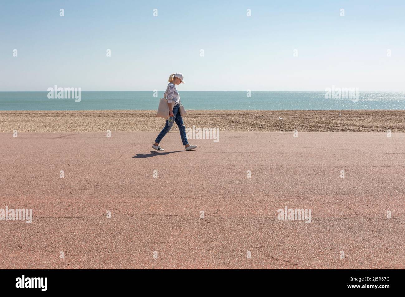 A solitary female walker on a promenade by a shingle beach and calm blue sea Stock Photo