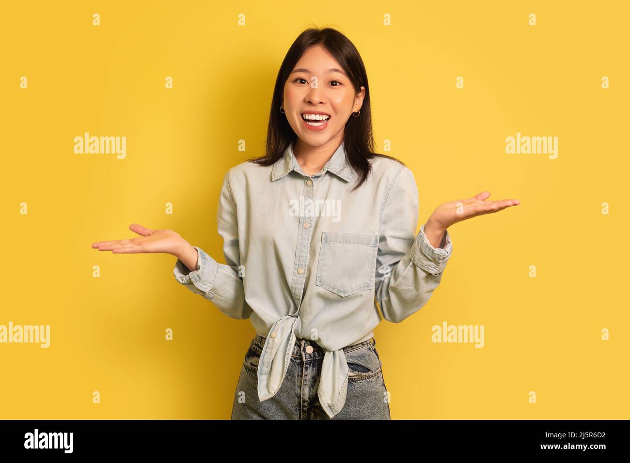 I don't know. Puzzled young Asian woman shrugging shoulders and smiling at camera on yellow studio background Stock Photo